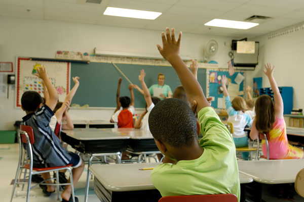 Teacher standing at the chalkboard and students raising their hands to answer a question