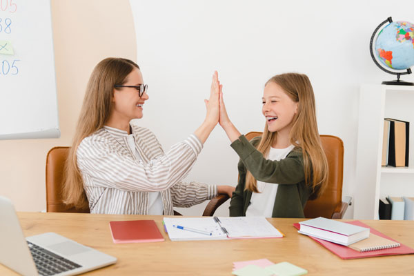 Teacher helping her student schoolgirl with schoolwork and giving high five at home school.